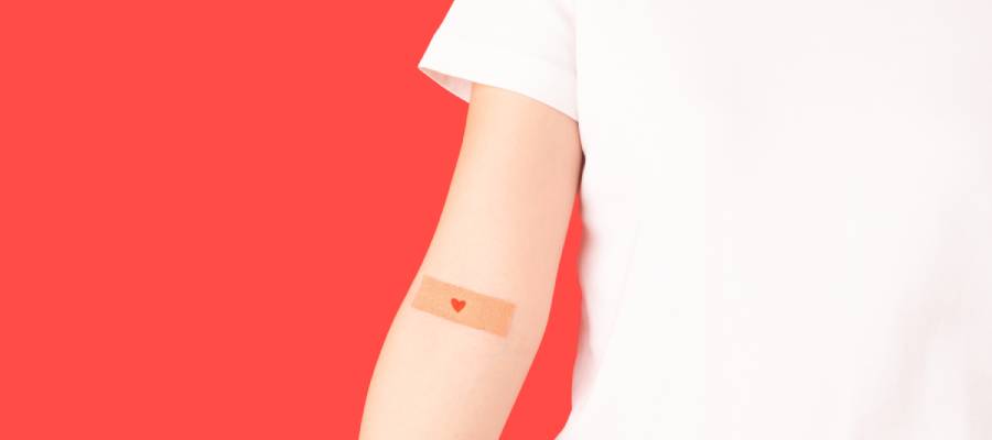 Kolunda küçük kırmızı kalp desenli bir yara bandı olan beyaz tişörtlü kişi, kan bağışı veya aşı sonrası görseli.