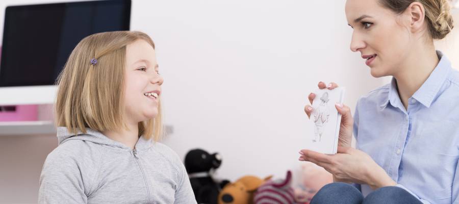 Bir çocuk ve bir yetişkin, bir odada birlikte oturuyor. Yetişkin, çocuğa bir figür çiziminin bulunduğu bir kart gösteriyor. Çocuk gülümsüyor ve dikkatle dinliyor. Arka planda oyuncaklar ve bir bilgisayar masası yer alıyor.