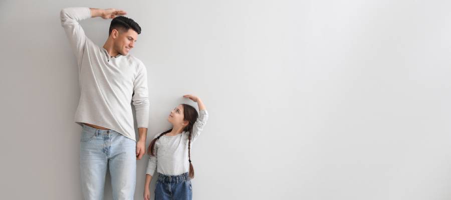 Bir baba ile küçük kızı, duvar önünde birbirlerinin boyunu ölçerken gülümsüyor. Baba ve çocuk ellerini başlarının üstüne koyarak karşılaştırma yapıyor. Aralarındaki boy farkı dikkat çekiyor.