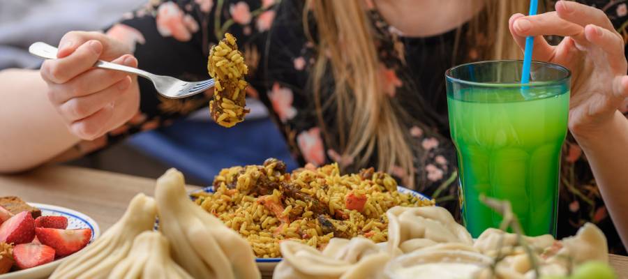 Bir kadın, önündeki pilav, mantı ve meyvelerden oluşan büyük porsiyonlu bir öğünü yerken yeşil bir içecek içiyor.