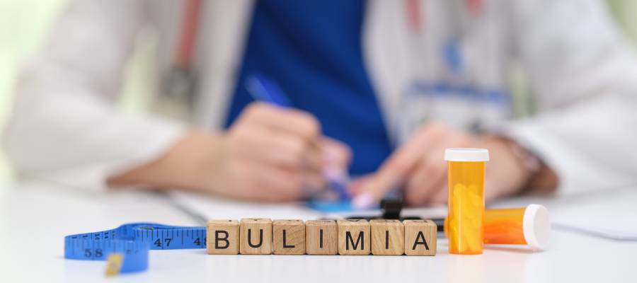 Bulimia yazılı tahta blokların önünde mezura, ilaç kutuları ve arka planda not alan bir sağlık çalışanı; bulimia nervozanın tıbbi ve psikolojik yönlerini simgeleyen bir görsel.