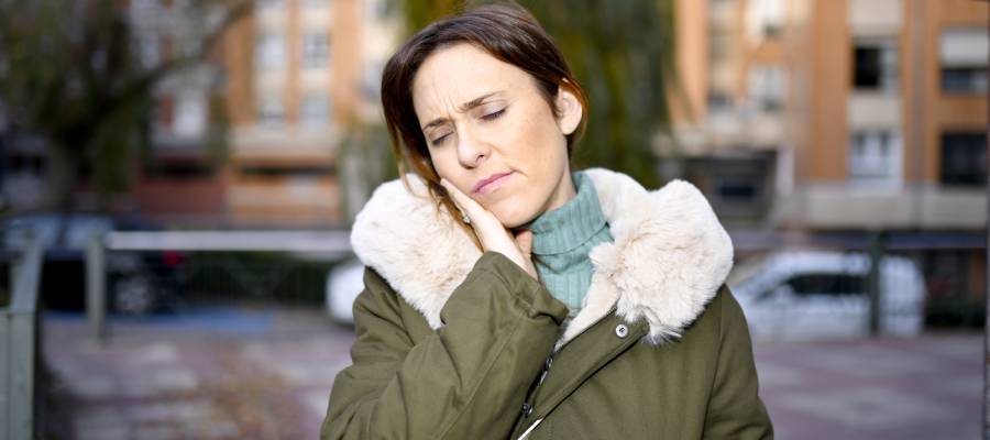 Dış mekânda yeşil mont giymiş bir kadın, başını hafif yana eğmiş ve eliyle çene eklemine dokunarak ağrılı bir ifade sergiliyor. Yüzündeki rahatsızlık ifadesi çene eklemi (temporomandibular eklem) ağrısını yansıtıyor