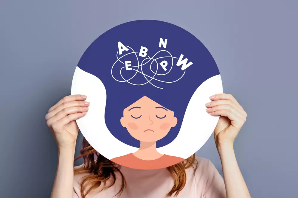 Disleksi yaşayan bir bireyi simgeleyen illüstrasyon: Bir kişi, kafa karışıklığını yansıtan karışık harfler (A, B, E, N, P, W) ile dolu bir daire şeklinde afişi yüzüne tutuyor.