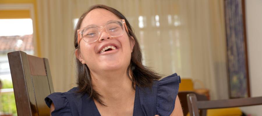 Gözlüklü, koyu saçlı ve koyu mavi elbise giymiş Down sendromlu genç bir kadın, içten bir kahkaha atarken sandalyesinde oturuyor.”