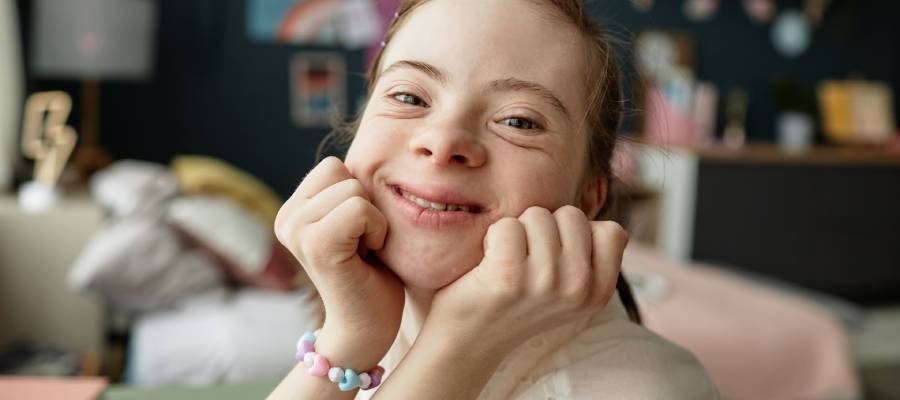Gülümseyen Down sendromlu genç bir kız, ellerini çenesine yaslamış şekilde kameraya bakıyor; arka planda yumuşak tonlarda dekore edilmiş bir oda bulunuyor.