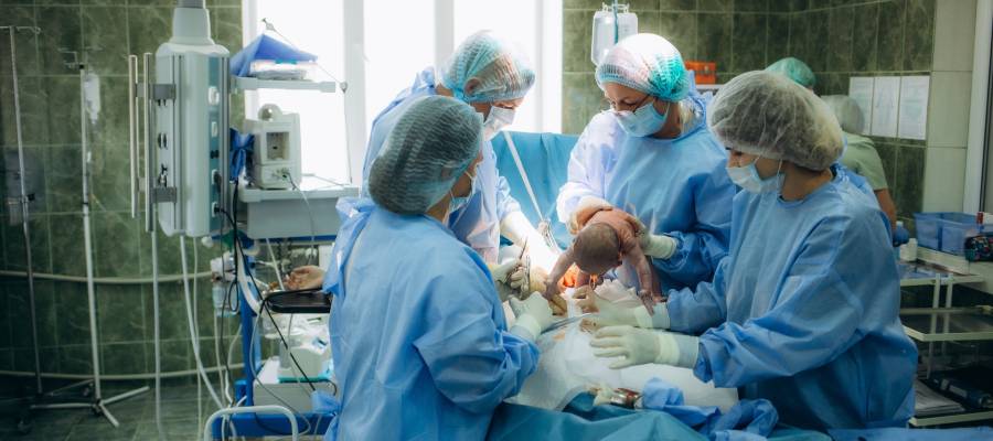 Doğumhanede sağlık çalışanları tarafından gerçekleştirilen vajinal doğum anı. Yeni doğan bebek annenin rahminden çıkarılırken görülüyor. Görsel, doğum sırasında uygulanan tıbbi müdahaleleri, özellikle epizyotomi işlemini (doğum kesisi) temsil etmektedir.