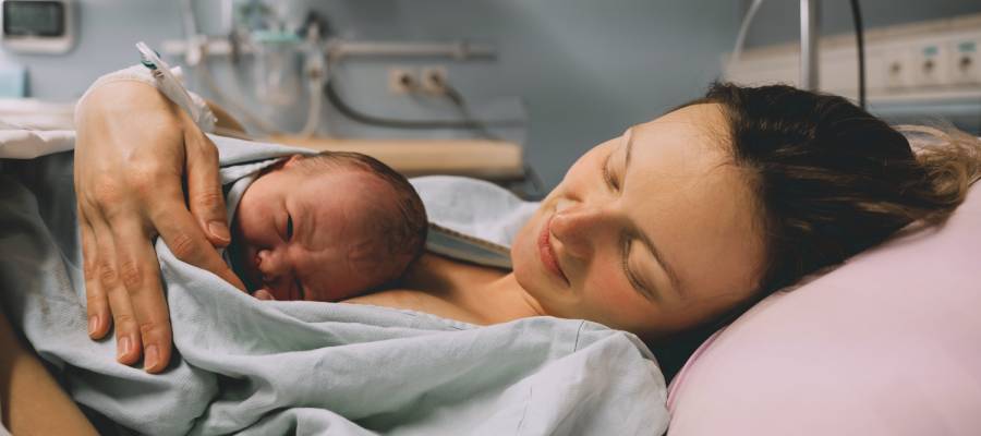 Doğum sonrası hastane yatağında yeni doğan bebeğini göğsünde tutan ve gülümseyen anne. Görsel, epizyotomi uygulamasının ardından yaşanan iyileşme sürecinde anne-bebek temasının önemini ve duygusal bağı simgelemektedir.