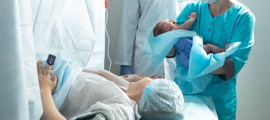 Sezaryen doğum sonrasında ameliyathanede yeni doğan bebeği tutan sağlık personeli ve perde arkasından bebeğine uzanan anne. Görsel, doğum şekline bakılmaksızın epizyotominin yalnızca vajinal doğumlarda uygulandığını ve farklı doğum senaryolarında müdahale biçimlerinin değiştiğini anlatmak için kullanılabilir.