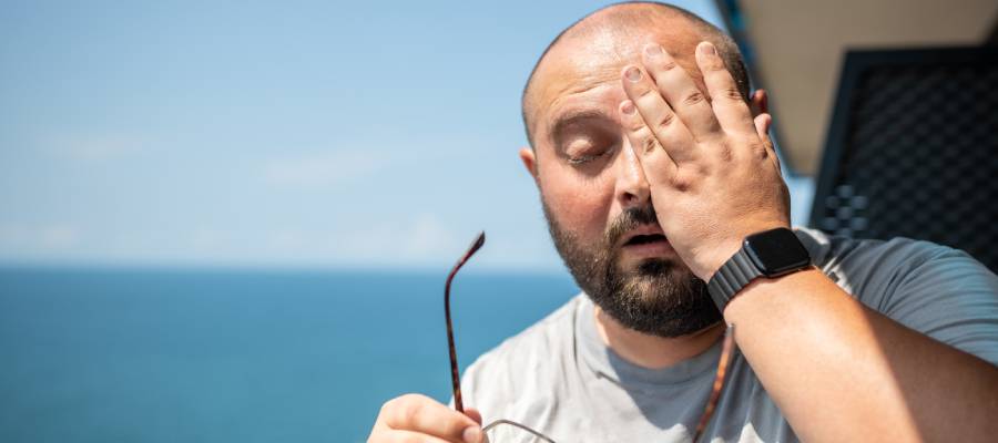 Gözlüklerini çıkarmış, gözünü ovuşturan ve yüzünde rahatsızlık ifadesi bulunan kel bir adam, deniz manzaralı açık bir alanda duruyor. Göz ağrısı veya ışığa hassasiyet gibi bir göz rahatsızlığı yaşıyor olabilir.