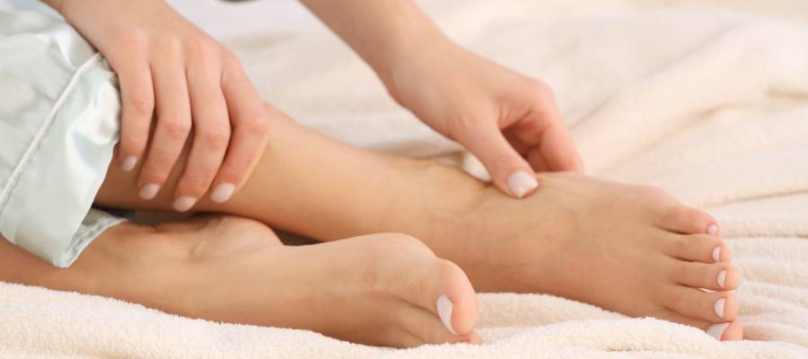 Bir kişi yatakta otururken ayak bileğini tutarak kaşıyor ya da inceliyor.