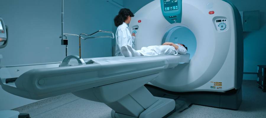 Bir hastane ortamında MR (Manyetik Rezonans) cihazına yatay pozisyonda yerleştirilmiş bir hasta ve cihazı kontrol eden beyaz önlüklü sağlık personeli.