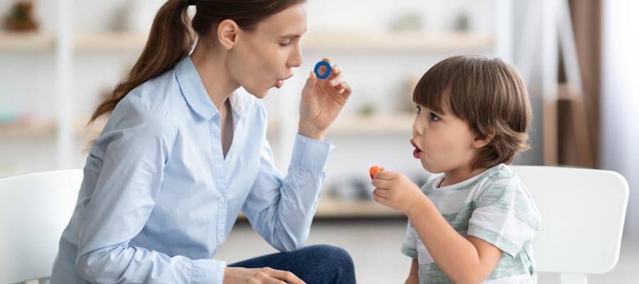 Bir kadın ve küçük bir erkek çocuk karşılıklı oturmuş, dudaklarını büzerek nefes üfleme egzersizi yapıyorlar. Kadının ve çocuğun ellerinde üfleme oyuncakları bulunuyor. Arka planda ev ortamı görülüyor.