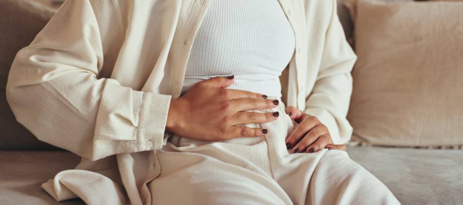 Karnını tutan kadın; mide bulantısı yaşayan bir kişi evde koltukta oturuyor.