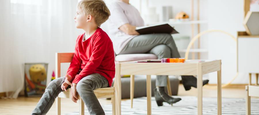 Kırmızı kazak giymiş küçük bir erkek çocuk, küçük bir sandalyede oturuyor ve yana doğru bakıyor. Arka planda bir uzman ya da terapist, dizlerinin üzerinde bir defterle oturmuş gözlem yapıyor. Ortam, bir değerlendirme veya terapi odasını andırıyor.