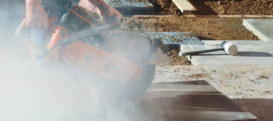 Bir işçinin beton veya taş kesme makinesiyle çalışırken yoğun toz bulutuna maruz kalması – silikozis riskini artıran tozlu çalışma ortamı.