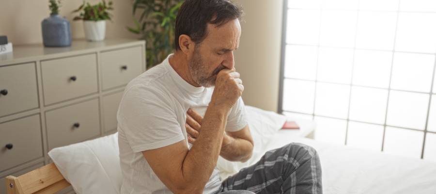 Yatakta otururken öksüren ve göğsünü tutan orta yaşlı bir adam – silikozis gibi kronik akciğer hastalıklarının tipik belirtilerinden biri olan nefes darlığı ve öksürükle mücadele ediyor.
