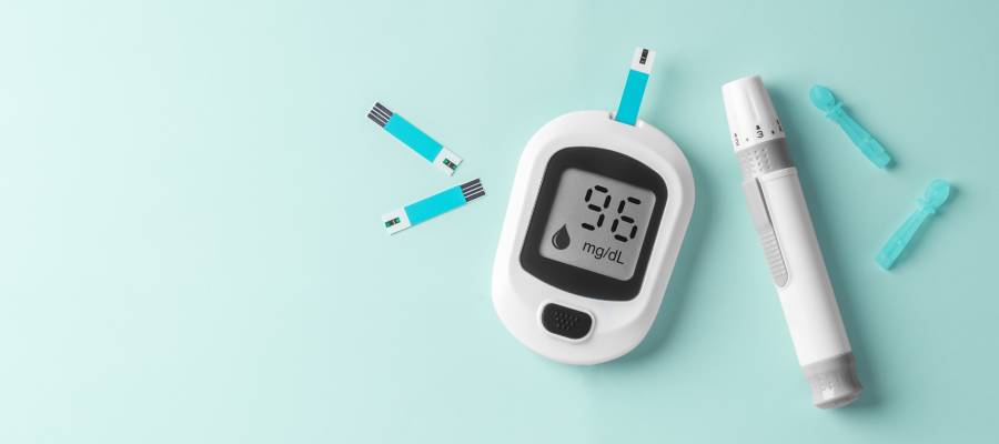 Bir kan şekeri ölçüm cihazı, test çubukları ve parmak delme kalemi, arka planda açık mavi zemin üzerinde. Cihaz ekranında 96 mg/dL kan şekeri seviyesi gösteriliyor.