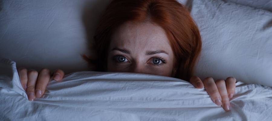 Gece uyanmış ve korkmuş görünen genç bir kadın yatakta yorganın altından gözleriyle endişeyle bakıyor.