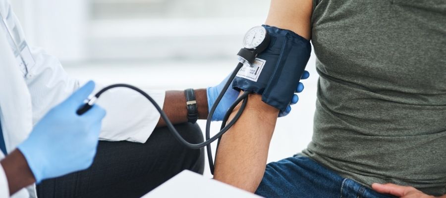 Bir sağlık çalışanı, glov giyerek oturan bir hastanın tansiyonunu mekanik tansiyon aletiyle ölçüyor.