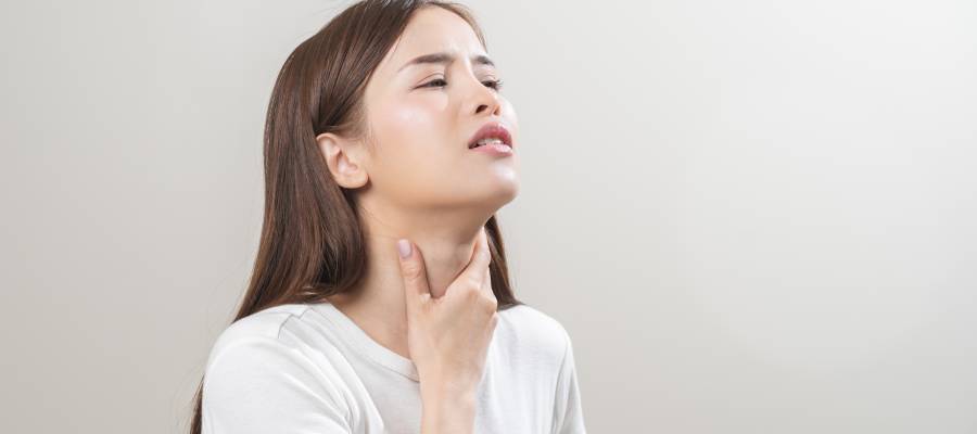 Boğazını tutarak rahatsızlık hissi yaşayan genç bir kadın; boğaz ağrısı belirtilerini gösteren beyaz arka planlı bir görsel.