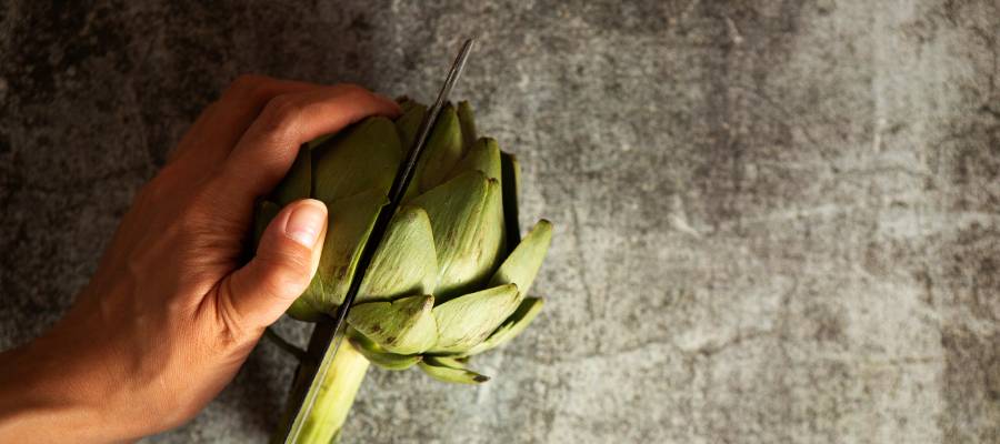 Koyu renkli tezgâh üzerinde bir kişinin eliyle tuttuğu enginar, keskin bir bıçakla ortadan ikiye kesilirken görülüyor.