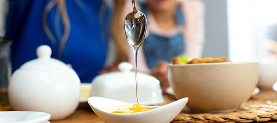 Bir kaşıktan beyaz bir kaseye bal dökülürken yakın çekim. Arka planda kahvaltı sofrası ve bulanık şekilde görünen insanlar bulunuyor.