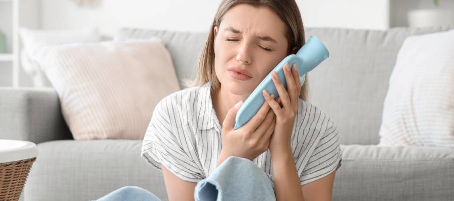 Evde koltuğun önünde oturan genç bir kadın, şiddetli diş ağrısı yaşıyormuş gibi yüzünü buruşturmuş ve mavi bir buz torbasını yanağına bastırıyor. Rahatsızlığı yüz ifadesinden belli oluyor. Arka planda yastıklı bir koltuk yer alıyor.