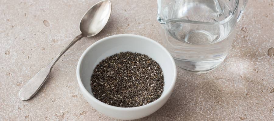 Beyaz bir kasede chia tohumu, yanında bir bardak su ve metal kaşık, açık renkli bir zemin üzerinde duruyor.