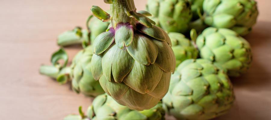 Ahşap zemin üzerinde taze ve yeşil yapraklarıyla dikkat çeken bütün enginarlar, önde bir tanesi net şekilde odaklanmış şekilde görülüyor.