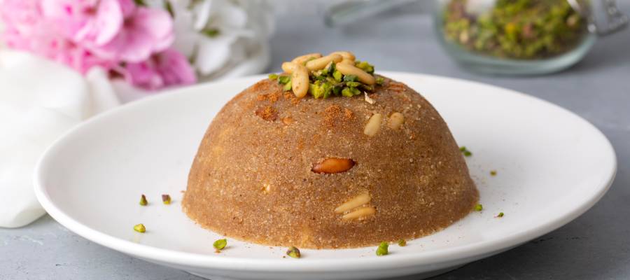 Yuvarlak şekilde kalıplanmış, fıstık ve çam fıstığıyla süslenmiş geleneksel Türk tatlısı irmik helvası, beyaz bir tabakta servis edilmiş.