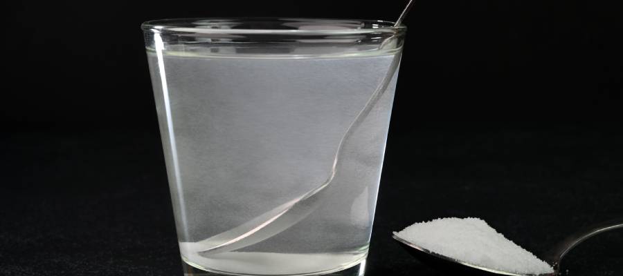 Bir bardak ılık tuzlu su ve yanında bir çay kaşığı dolusu tuz içeren metal kaşık; boğaz ağrısı için gargara hazırlığında sade, koyu arka planlı bir görüntü.