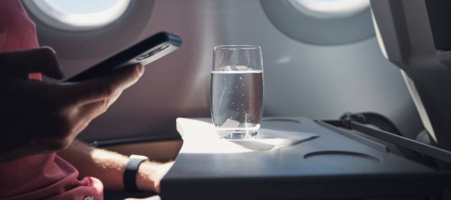 Uçakta pencere kenarında oturan bir yolcu, katlanır masa üzerinde duran bir bardak suya bakarken telefonunu tutuyor.