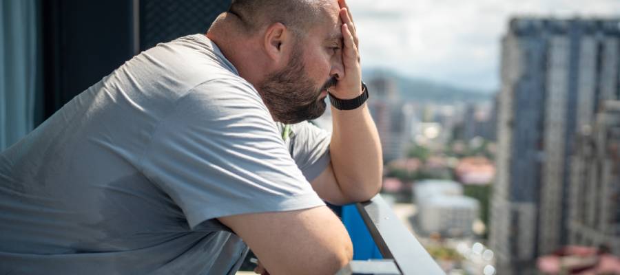 "Balkonda duran, yüzünü eliyle kapatan ve sırtı terden ıslanmış bir adam, sıcak hava veya stres kaynaklı aşırı terleme (hiperhidroz) yaşıyor.