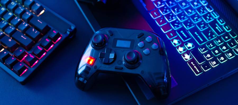 Renkli ışıklarla aydınlatılmış bir oyun ortamında, parlak RGB klavyeli bir bilgisayar ve onun yanında bulunan oyun kolu (gamepad). Görsel, dijital oyunların cezbedici estetiğini ve oyun bağımlılığının dikkat çeken görsel yanını yansıtıyor.