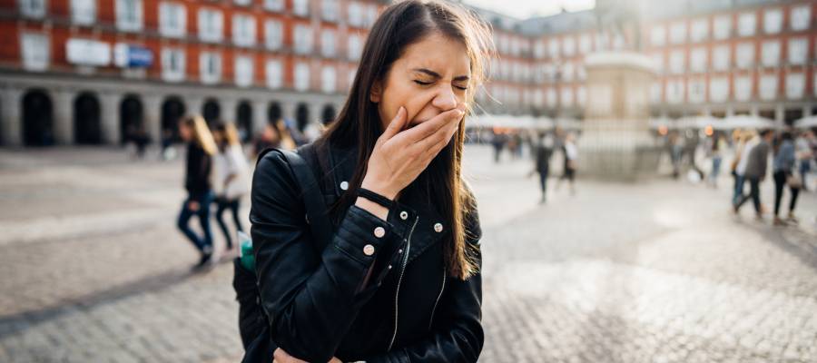 Tarihi bir şehir meydanında gezen genç bir kadın esnerken yorgun görünüyor; arka planda yürüyen insanlar ve klasik mimarili binalar bulunuyor