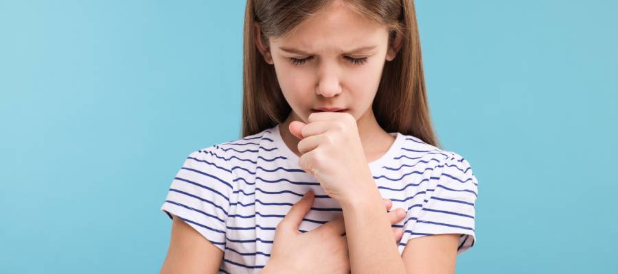 Göğsünü tutarak öksüren üzgün görünümlü bir kız çocuğu, mavi arka plan önünde.