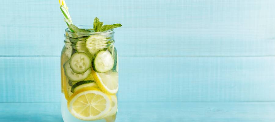 Bir mason kavanoz içinde dilimlenmiş limon, salatalık ve nane yapraklarıyla hazırlanmış serinletici bir detoks suyu. Kavanozda renkli bir pipet yer almakta ve arka planda açık mavi ahşap bir zemin bulunmakta.