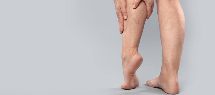 Gri arka planda ayakta duran bir kişinin alt bacağını tutması; bacakta damar belirginliği, şişlik veya varis sorunu belirtisi.